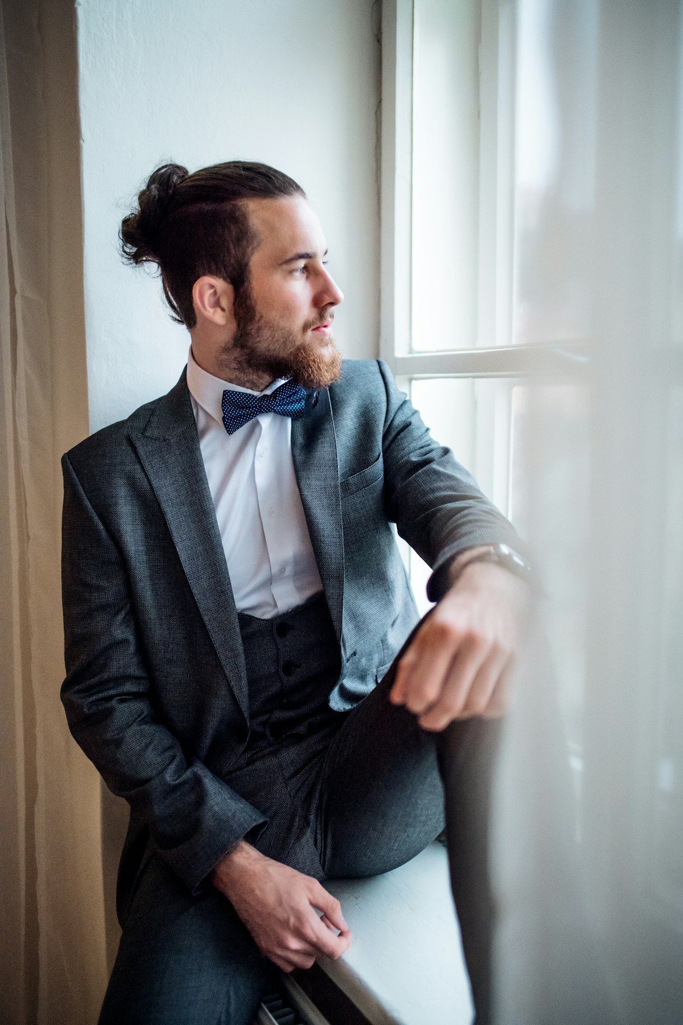 A handsome hipster young man with formal suit sitting on a window sill on an indoor party.